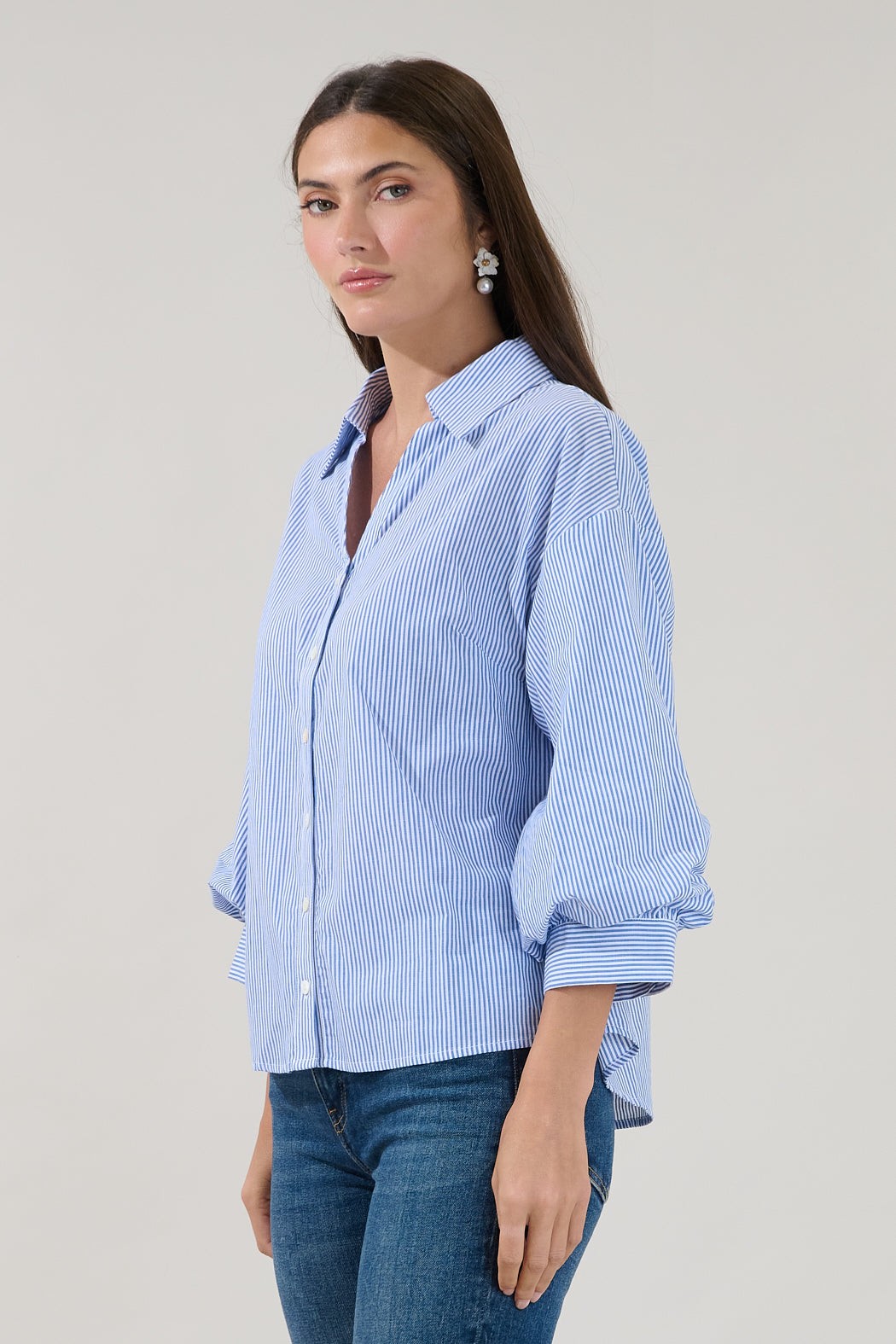 A woman with long brown hair wears the Sugarlips Chalet Button Down Stripe Top, featuring blue and white stripes and rolled-up sleeves, paired with blue jeans as she stands against a plain light background.