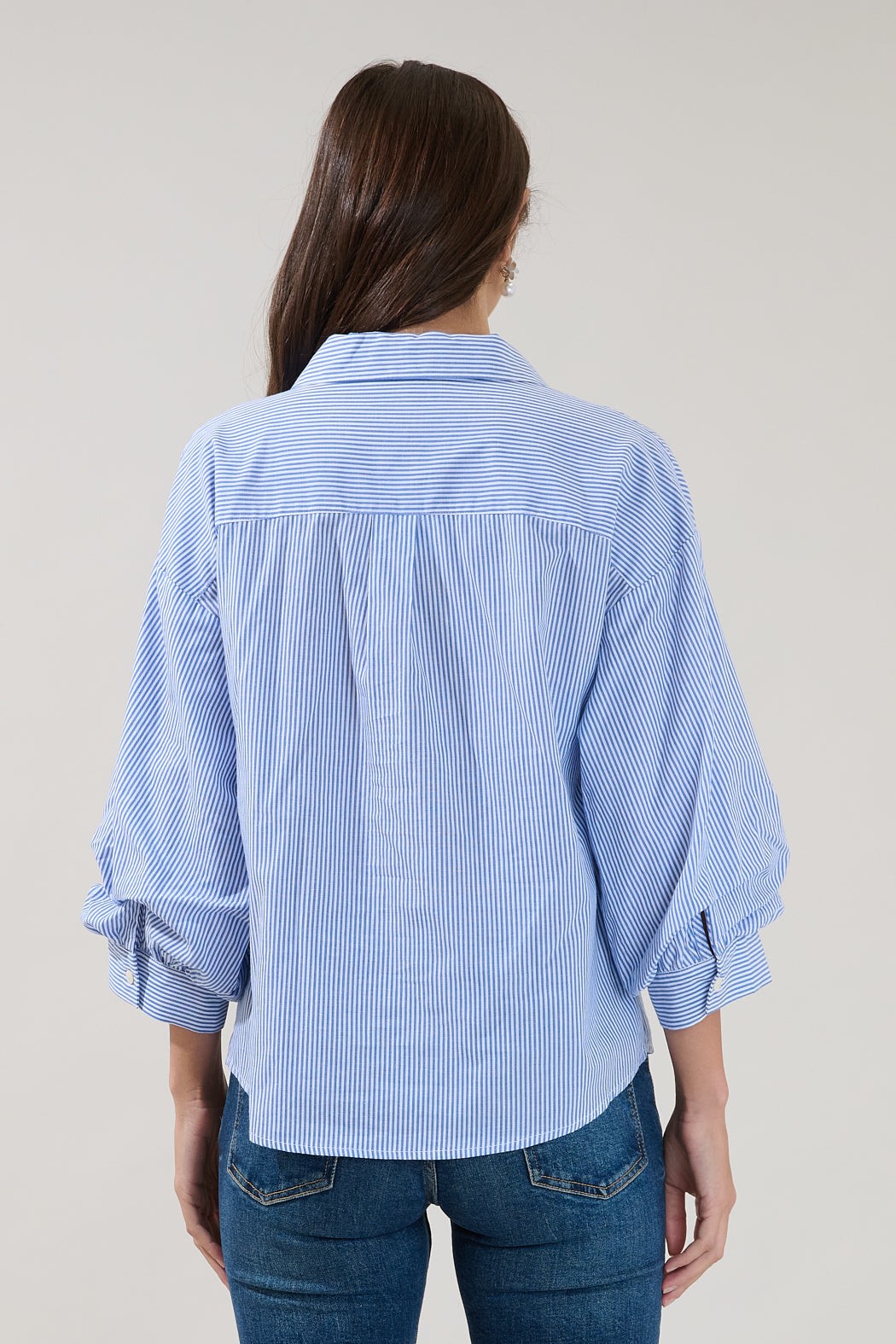 A person with long brown hair, seen from behind, wears the Sugarlips Chalet Button Down Stripe Top by Sugarlips—with blue and white stripes and rolled sleeves—paired with blue jeans against a plain background.