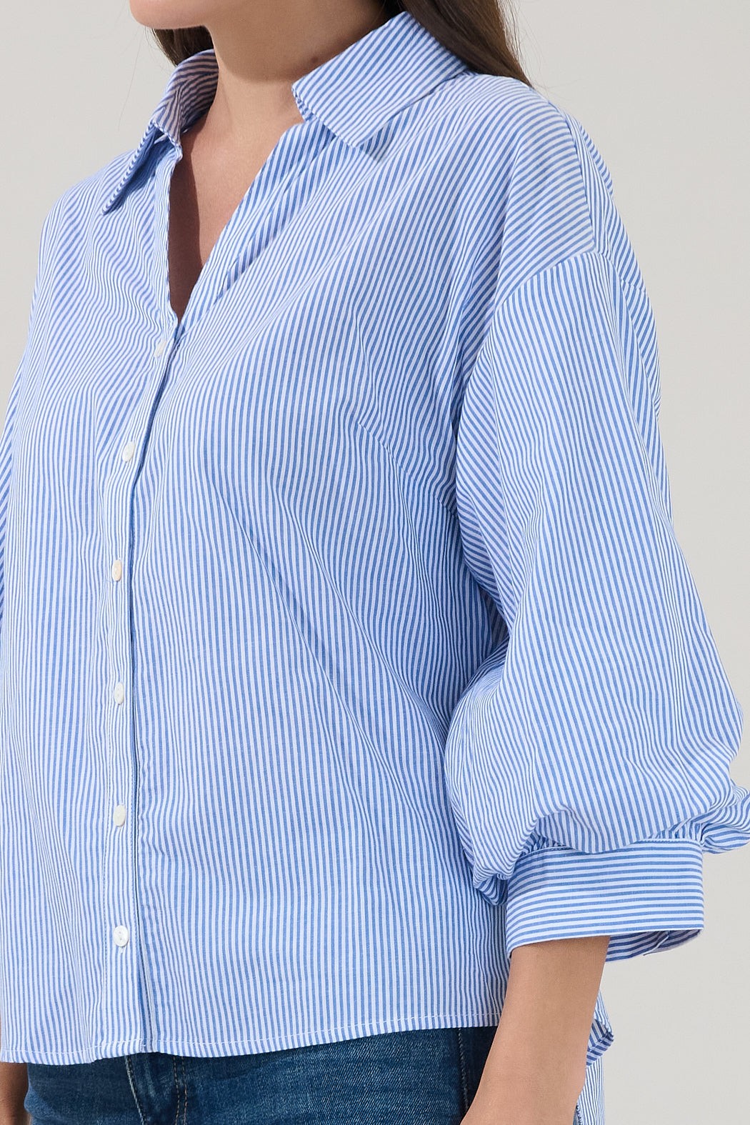 A person wearing the Sugarlips Chalet Button Down Stripe Top by Sugarlips, featuring blue and white vertical stripes with rolled-up sleeves, paired with blue jeans and standing against a plain background.