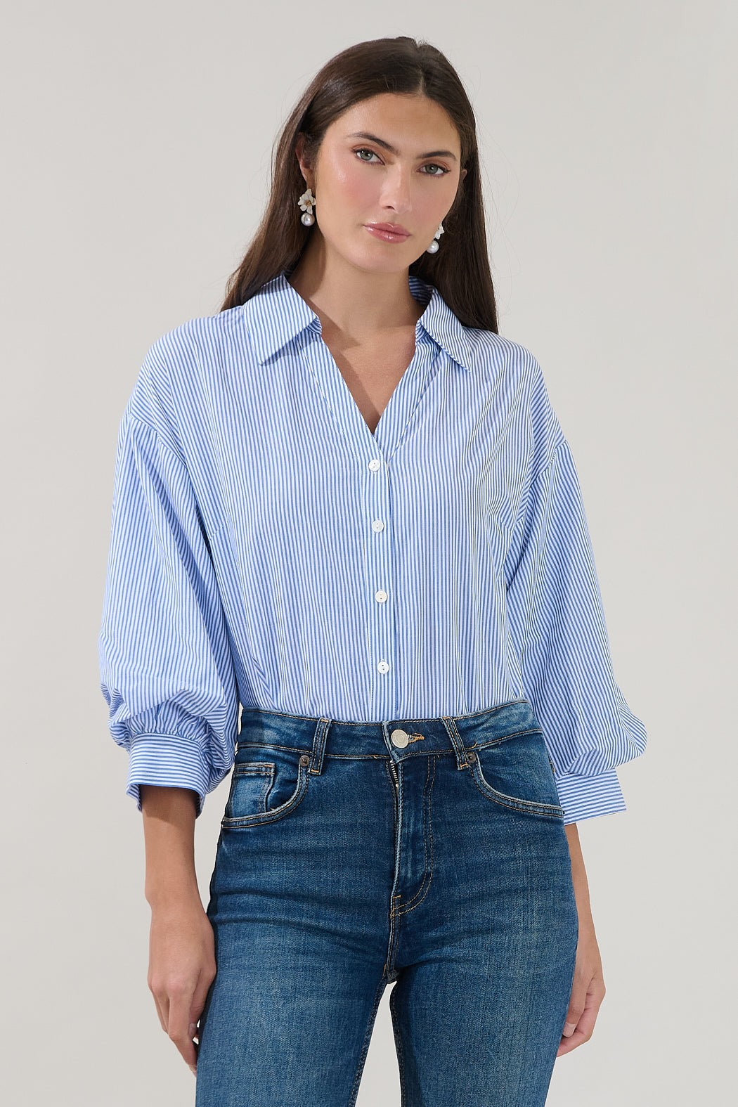A woman wears the Sugarlips Chalet Button Down Stripe Top with rolled sleeves and blue jeans, standing against a plain light background.