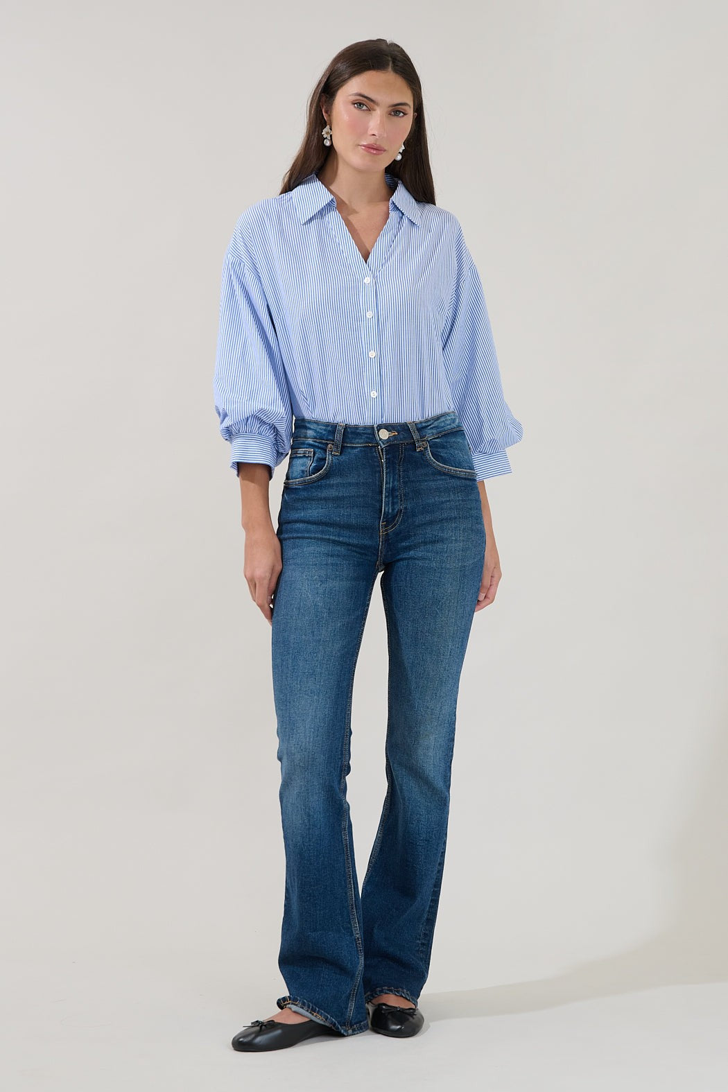A woman stands in front of a plain background wearing the Sugarlips Chalet Button Down Stripe Top, styled with blue jeans and black flats, her hands relaxed by her sides.