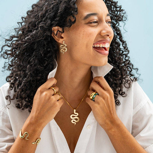 INK + ALLOY Kara Snake Post Earrings in brass worn on model with curly hair against blue background