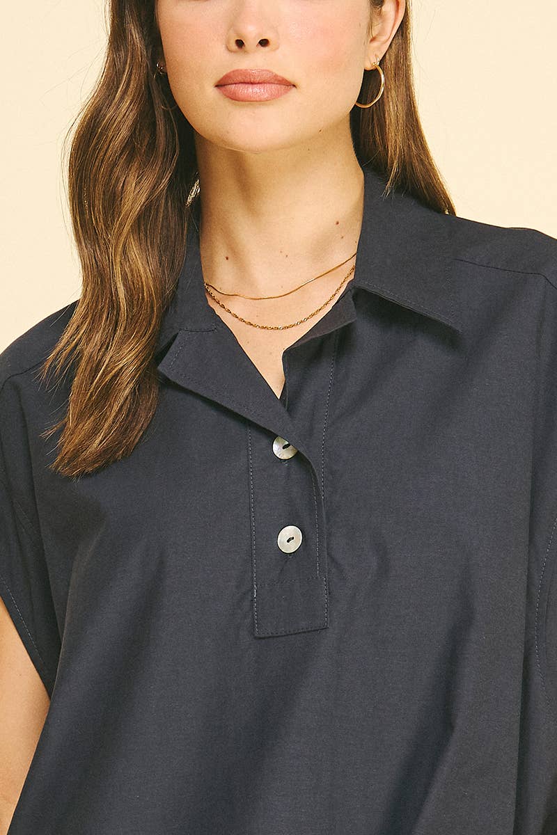 A woman wears the PINCH Solid Cap Sleeve Blouse Top by PINCH, featuring a navy color, classic collar, and three-button design. She accessorizes with gold hoop earrings and layered gold necklaces. Her wavy brown hair is styled loosely, set against a light beige background.