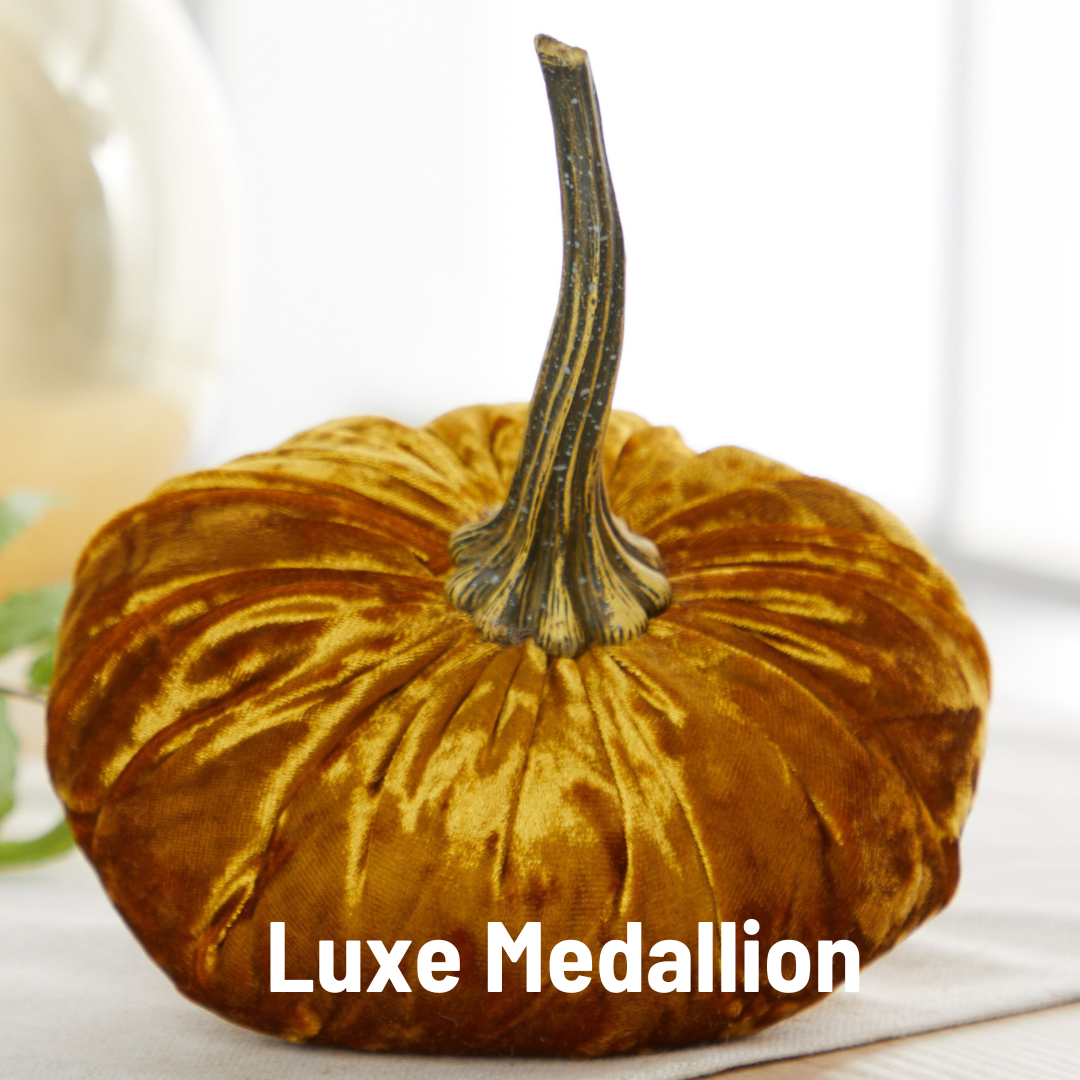 A plush velvet pumpkin crafted from lustrous golden-brown fabric with a lifelike dark stem. This handcrafted décor piece by Your Heart’s Content—named “Pumpkin Large Velvet”—is perfect for fall mantel displays or as an elegant Halloween table centerpiece, bringing a touch of luxury to any space. The softly blurred background highlights the image text: “Luxe Medallion.”.