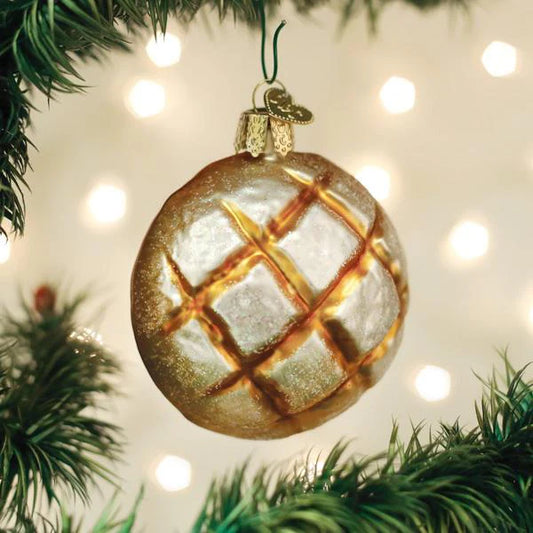 An Old World Christmas Sourdough Bread Ornament from Old World Christmas, shaped and painted like a golden-brown loaf with a rustic crust, hangs among green tree branches, evoking memories of holiday baking as soft, blurry lights twinkle in the background.