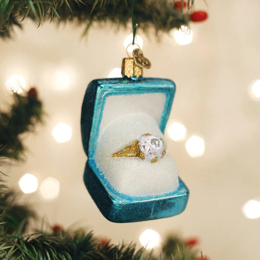 The Old World Christmas Something Blue Ring Box Ornament by Old World Christmas, featuring a charming blue ring box with a gold ring inside, hangs from a branch and evokes the magic of a bride’s wedding day among softly blurred twinkling white lights in the background.