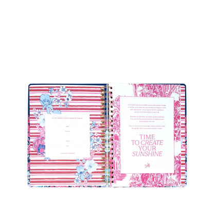 An open spiral-bound planner, the Lilly Pulitzer by Lifeguard Press - Jumbo 17 Month Agenda in "In All Her Glory," displays pink and white striped pages adorned with floral designs. On the right page, vibrant text reads "TIME TO CREATE YOUR SUNSHINE." Inspired by classic Lilly Pulitzer style, this hardcover agenda features a left page with spaces for personal information—ideal for keeping organized during your 2026-2027 planning.