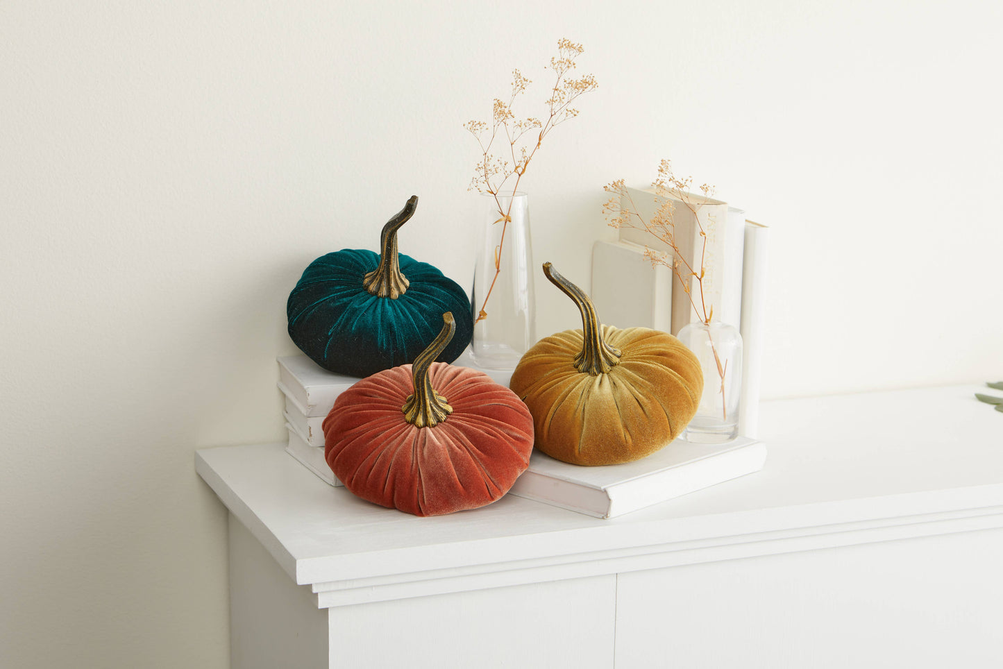 Three large velvet pumpkins by Your Heart’s Content add a handcrafted touch in rich teal, orange, and yellow hues, arranged atop stacked white books near glass vases of dried flowers—a warm display ideal for your Thanksgiving centerpiece or fall mantel decor.