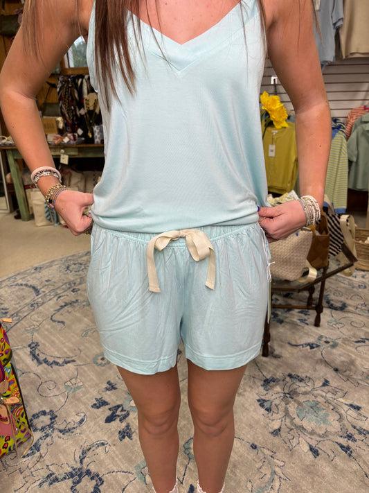 A woman is wearing the Boody Goodnight Sleep Shorts in Seafoam by Boody, paired with a light blue sleeveless top. She stands indoors holding the sides of her shorts, with patterned carpet and clothing racks visible in the background.