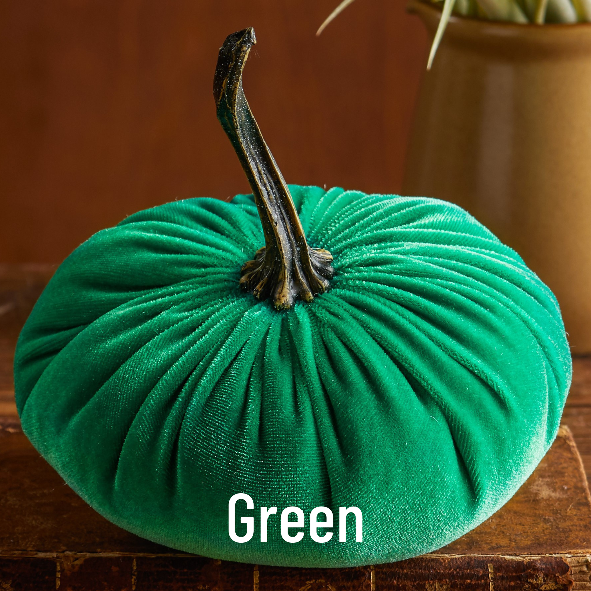 A large, plush green velvet pumpkin from Your Heart’s Content rests on a wooden surface, its dark curved stem adding a touch of elegance. This handcrafted fall mantel decor makes a charming addition to your Thanksgiving or Halloween table. The word “Green” appears at the bottom of the image.