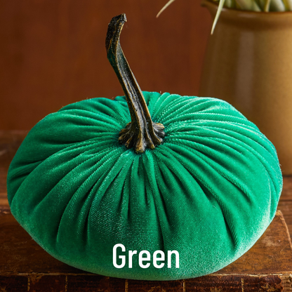 A large, plush green velvet pumpkin from Your Heart’s Content rests on a wooden surface, its dark curved stem adding a touch of elegance. This handcrafted fall mantel decor makes a charming addition to your Thanksgiving or Halloween table. The word “Green” appears at the bottom of the image.