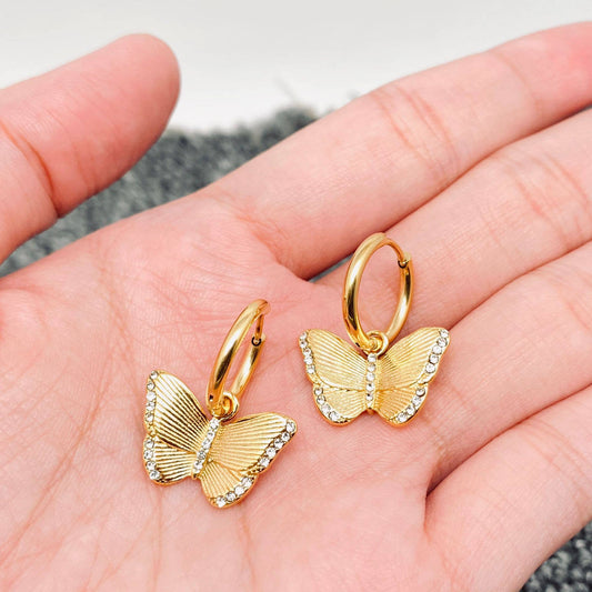 A hand displays the Mio Queena - Butterfly Gold Plated Huggie Earrings by Mio Queena, each featuring a charming butterfly adorned with sparkling clear stones along the edges. These stunning 18K gold plated huggie earrings are crafted from stainless steel, providing both elegance and durability as hypoallergenic jewelry.