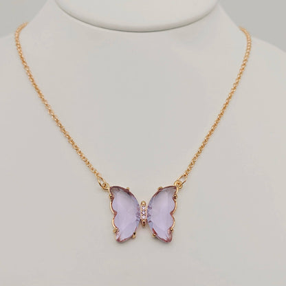 The Mio Queena - Multicolor Glass Butterfly Pendant Necklace by Mio Queena features a gold chain with a vibrant purple butterfly pendant, highlighted by a row of sparkling clear stones at its center. Displayed elegantly on a white jewelry bust, this hypoallergenic piece is both fashionable and gentle on sensitive skin.