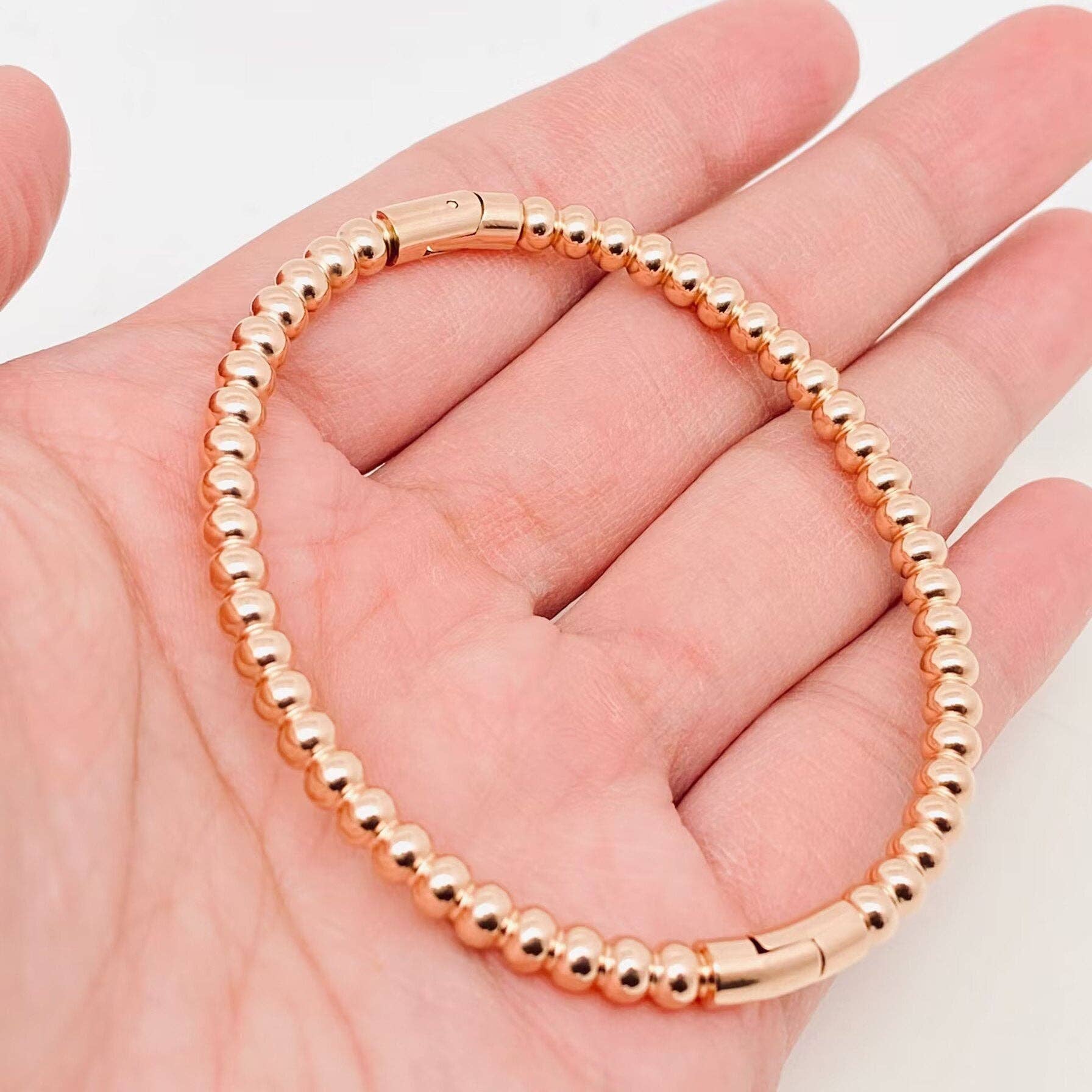 A hand displays the Mio Queena - 4mm Beads Gold Plated Stainless Steel Snap Bangle, featuring small round gold-plated beads and a secure snap clasp, set against a plain background—an elegant gift option from Mio Queena.