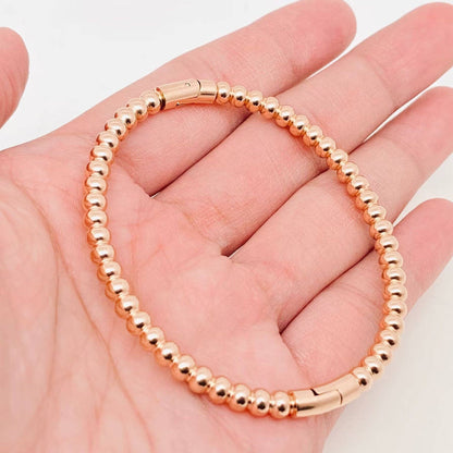 A hand displays the Mio Queena - 4mm Beads Gold Plated Stainless Steel Snap Bangle, featuring small round gold-plated beads and a secure snap clasp, set against a plain background—an elegant gift option from Mio Queena.