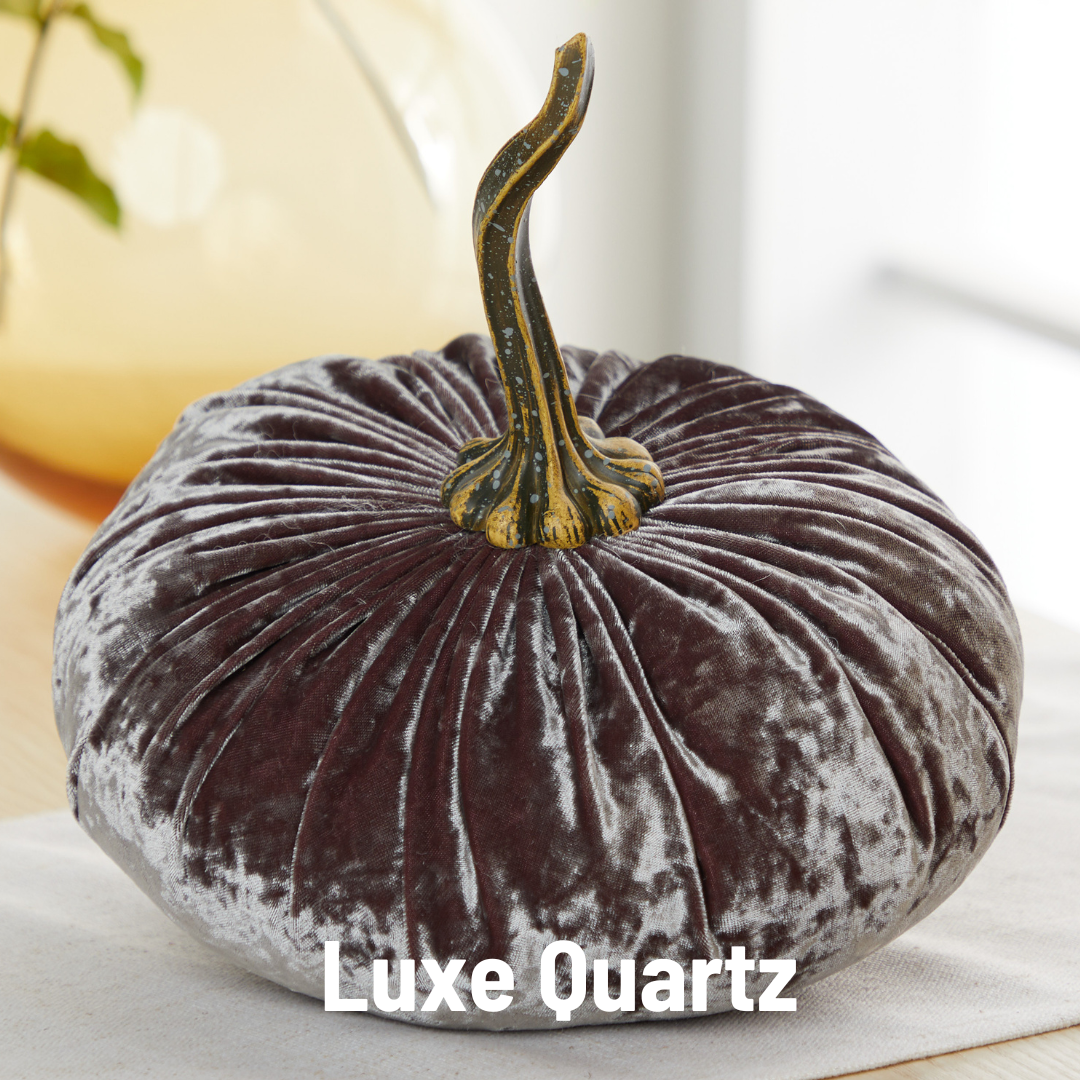 A silvery-gray, extra large velvet pumpkin from Your Heart’s Content features a gold and green stem and sits elegantly atop a table, making it an ideal choice for sophisticated holiday mantel décor. At the bottom, “Luxe Quartz” is displayed in white text.