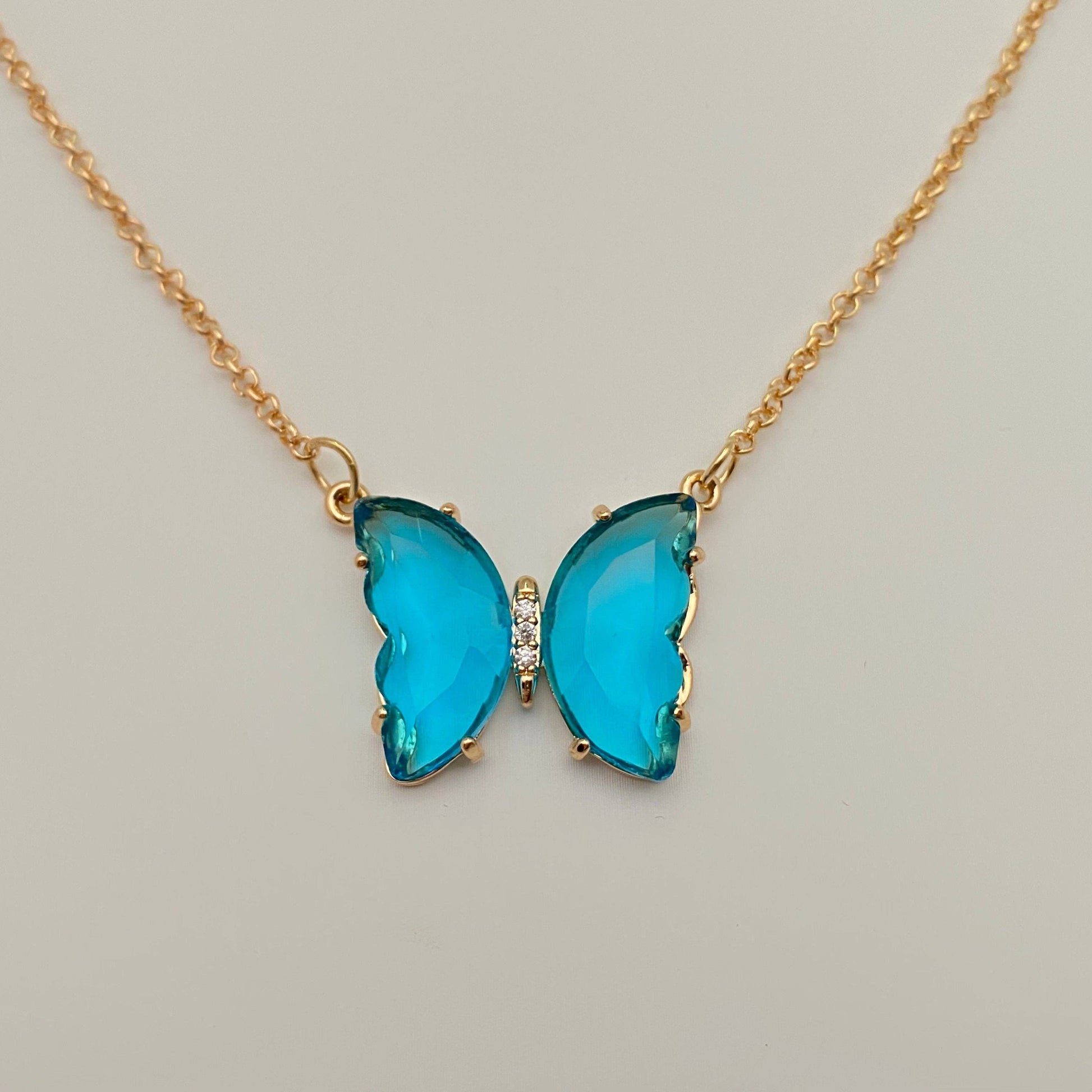 The Mio Queena - Multicolor Glass Butterfly Pendant Necklace by Mio Queena features a delicate butterfly-shaped pendant, adorned with two blue glass wings and a small row of clear stones at the center. Crafted as hypoallergenic jewelry, this elegant piece is showcased against a plain, light-colored background.