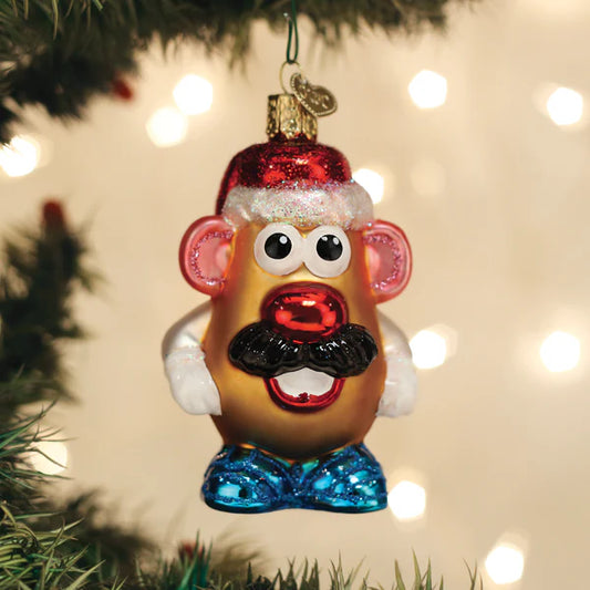 The Old World Christmas Mr. Potato Head Ornament by Old World Christmas is a festive decoration shaped like the classic toy, complete with a red Santa hat, black mustache, blue shoes, and googly eyes, hanging from a tree branch with holiday lights twinkling in the background.