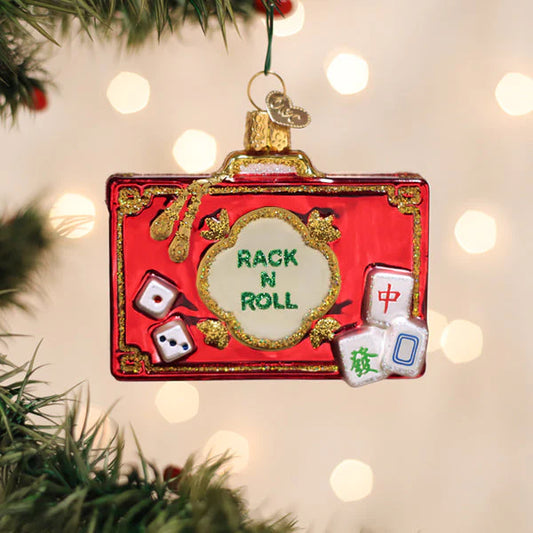 An Old World Christmas Rack N Roll Mahjong Ornament by Old World Christmas, designed in the shape of a red suitcase with gold trim, hangs on a festive tree. Featuring dice and mahjong tiles with “RACK N ROLL” displayed across the front, this ornament celebrates the classic game amid softly blurred holiday lights in the background.