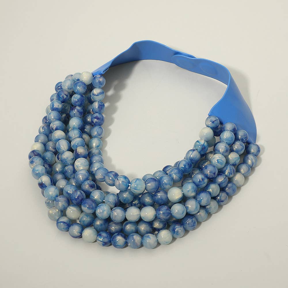 The Sensibling Corp. - Marbled Ball Beaded Multi Layered Faux Leather Magnetic Collar Necklace, by Sensibling Corp., is displayed on a plain white background. This eye-catching statement piece features multiple layered strands with round blue and white marbled beads, completed with a wide, glossy blue magnetic closure for secure and stylish fastening.