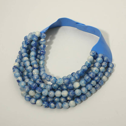 The Sensibling Corp. - Marbled Ball Beaded Multi Layered Faux Leather Magnetic Collar Necklace, by Sensibling Corp., is displayed on a plain white background. This eye-catching statement piece features multiple layered strands with round blue and white marbled beads, completed with a wide, glossy blue magnetic closure for secure and stylish fastening.