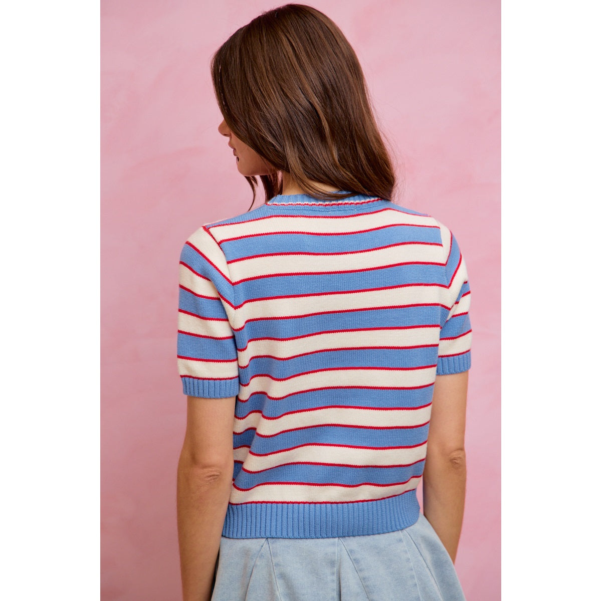 A woman with long brown hair, seen from behind, wears the So Me Stripe Knit Crew Neck Short Sleeve Sweater Top by So Me—a chic knit sweater featuring red and white horizontal stripes on a blue background—paired with a light blue skirt, set against a soft pink backdrop.