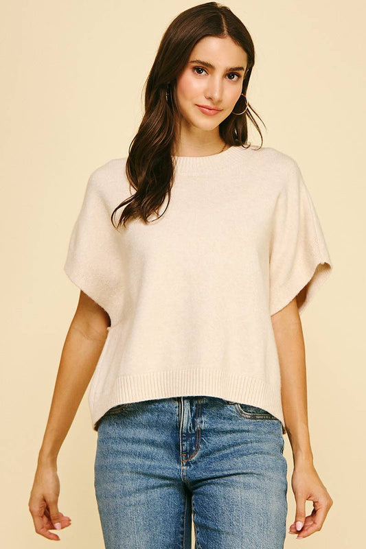 A woman with long brown hair is wearing the PINCH - DROPPED SHOULDER SWEATER TOP, a loose fit, short-sleeve cream sweater with a round neck and dropped shoulders from PINCH. She pairs it with blue jeans and stands against a beige background, looking at the camera with a slight smile.