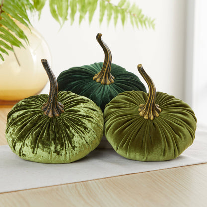 Three plush, velvet pumpkins in assorted green hues from Your Heart’s Content—featuring realistic brown stems—are arranged on a light wooden surface. The handcrafted “Pumpkin Large Velvet” decor by Your Heart’s Content makes an elegant Thanksgiving centerpiece; in the softly blurred background, a leafy fern and a yellow vase add to the cozy fall atmosphere.