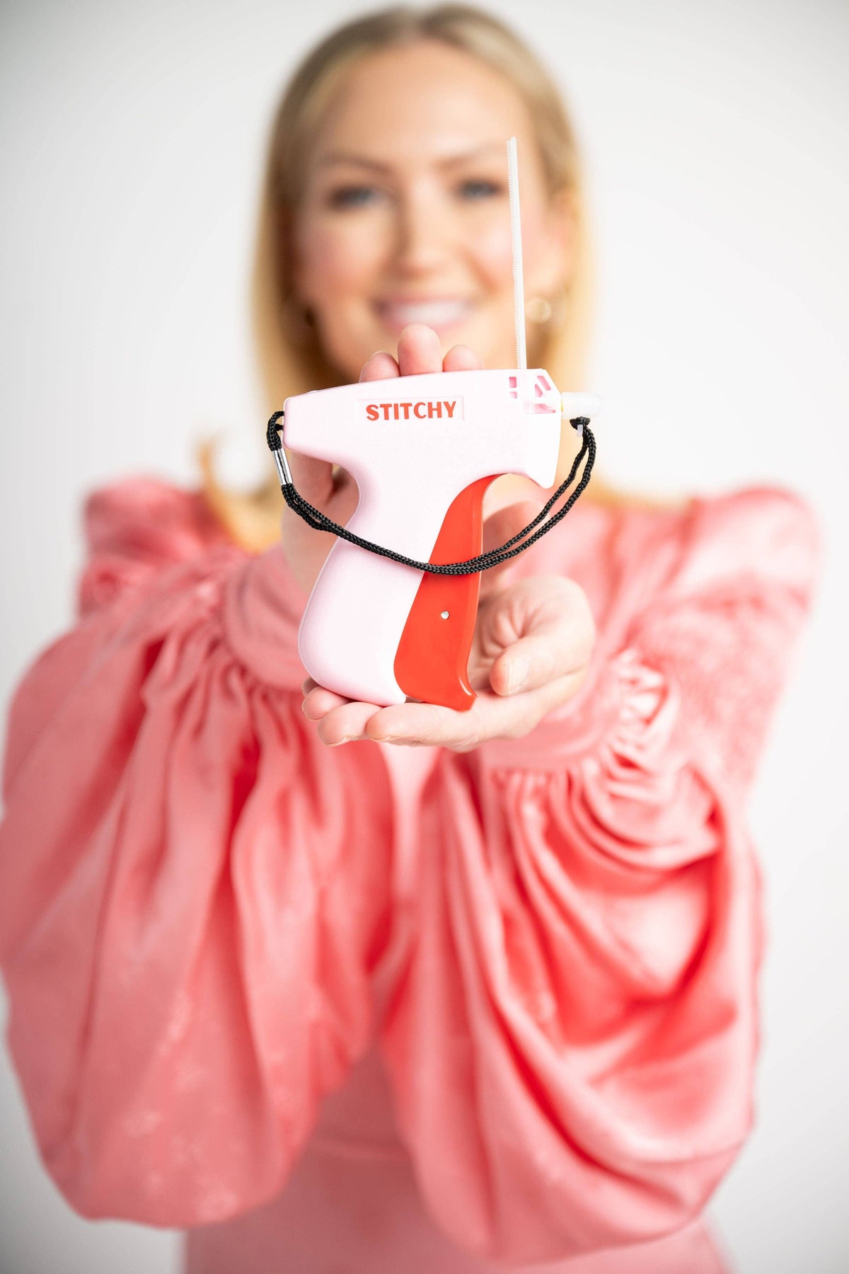 Woman in coral dress holding pink and red Tucky Stitchy tag gun toward camera