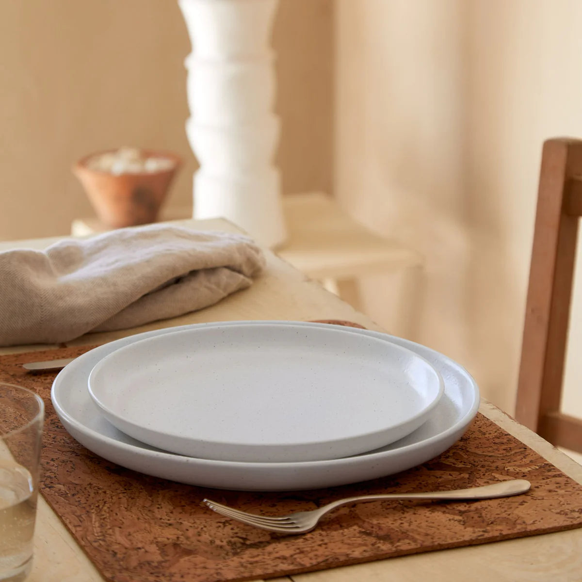 Two Costa Nova Casafina “Pacifica - White” dinner plates are stacked on a cork placemat alongside a fork atop a wooden table. A beige napkin, glass of water, and neutral decor complete the softly lit setting.