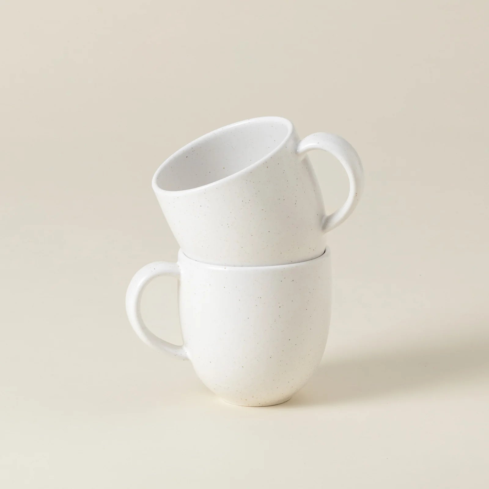 Two Casafina “Pacifica - White” mugs by Casafina are stacked, with one mug resting inside and slightly tilted on top of the other, set against a plain beige background.