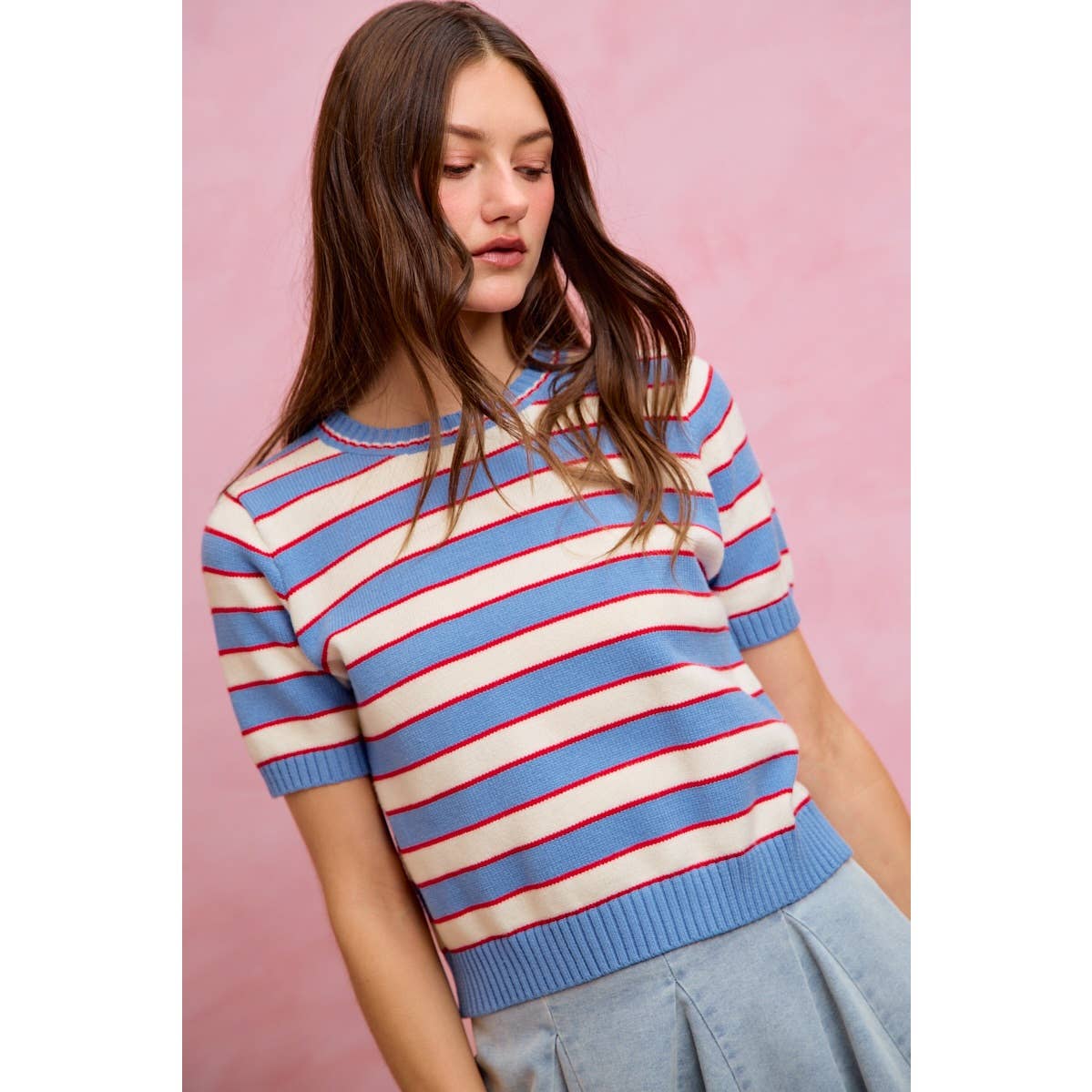 A woman with long brown hair is wearing the So Me STRIPE KNIT CREW NECK SHORT SLEEVES SWEATER TOP, featuring blue and white stripes with red accents, paired with a light blue skirt, while standing in front of a pink background.
