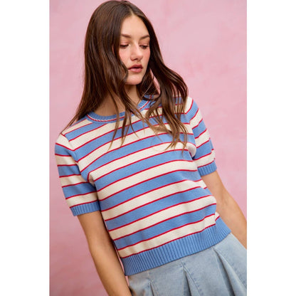 A woman with long brown hair is wearing the So Me STRIPE KNIT CREW NECK SHORT SLEEVES SWEATER TOP, featuring blue and white stripes with red accents, paired with a light blue skirt, while standing in front of a pink background.