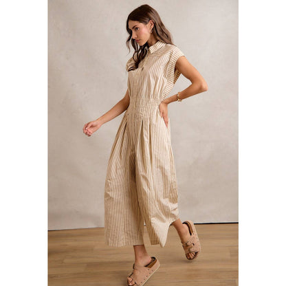 A woman models the So Me - SLEEVELESS WIDE LEG STRIPE JUMPSUIT, featuring beige and white stripes and a wide-leg silhouette, paired with tan sandals on a wooden floor against a light gray background.