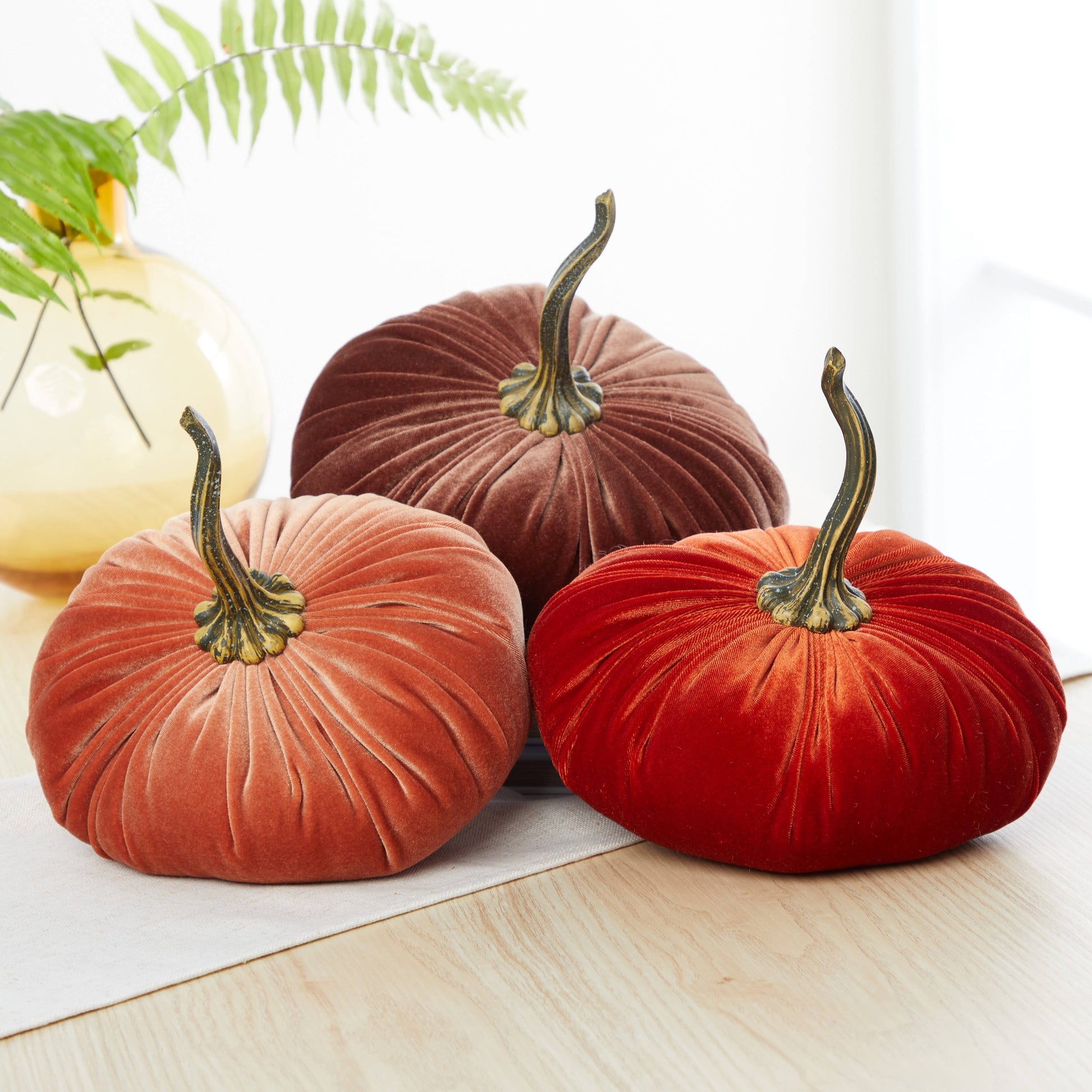 An extra large velvet pumpkin from Your Heart’s Content, in rich fall colors, sits as a striking accent on a wooden table with a cream runner—perfect for Thanksgiving decor—while a yellow vase filled with green leaves completes the cozy autumn setting.