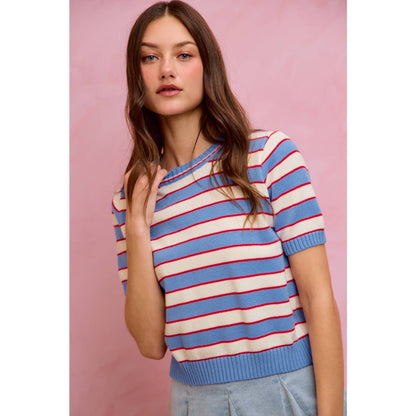 A woman with long brown hair is wearing the So Me STRIPE KNIT CREW NECK SHORT SLEEVES SWEATER TOP, a trendy blue and white striped knit sweater with red lines by So Me, paired with a light blue skirt. She stands in front of a pink background.