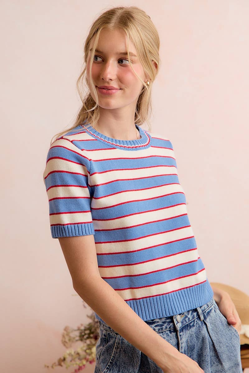 A woman with blonde hair wears the So Me STRIPE KNIT CREW NECK SHORT SLEEVES SWEATER TOP—featuring light blue, white, and red stripes—paired with high-waisted blue jeans, standing against a light pink background.