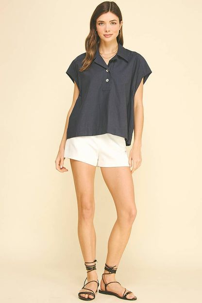 A woman stands against a beige background wearing the PINCH - SOLID CAP SLEEVE BLOUSE TOP by PINCH, styled with classic white shorts and black strappy sandals. She smiles slightly, with one hand resting by her side.