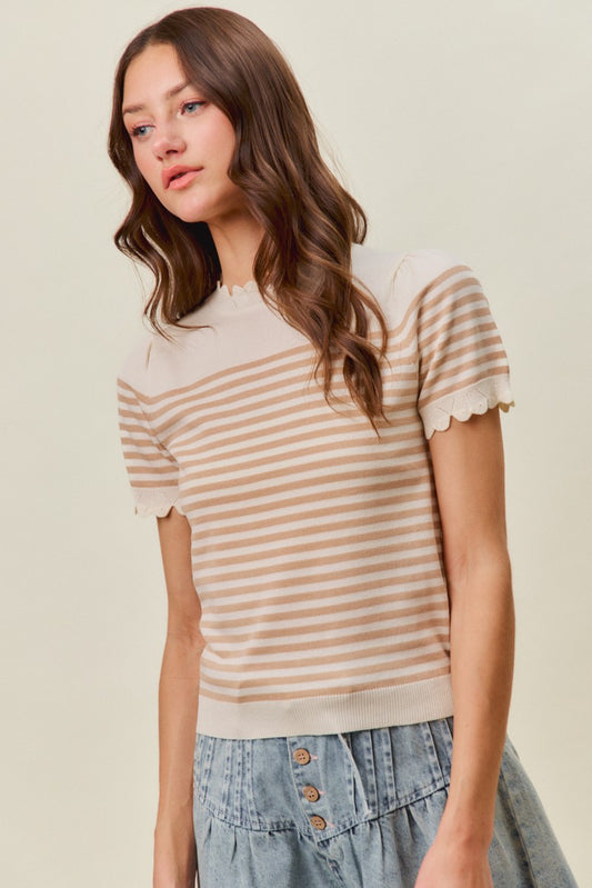 A woman with long wavy brown hair stands against a light neutral background wearing the So Me Striped Lightweight Sweater by So Me, featuring beige and white stripes and short sleeves, paired with a light blue button-front denim skirt.