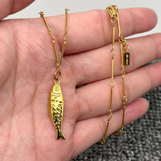 A hand holds the Mio Queena - 18K Gold Plated Stainless Steel Fish Necklace, featuring a fish-shaped pendant and an elongated, linked chain. The necklace rests against a gray textured background.