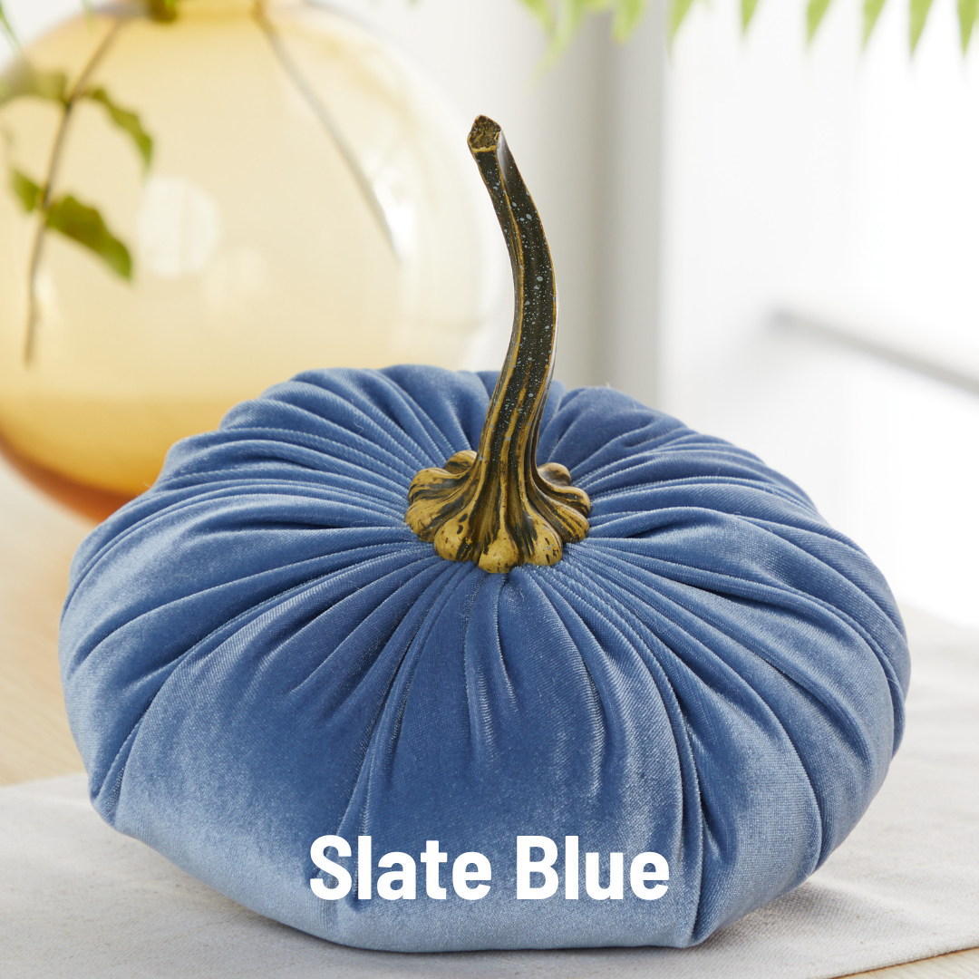 An extra-large velvet pumpkin from Your Heart’s Content, in a refined slate blue hue with a lifelike brown and green stem, sits elegantly on a table—an ideal accent for wedding centerpieces or Thanksgiving table décor. “Slate Blue” is written across the front of the plush pumpkin, while a blurred glass vase with a plant appears in the background.
