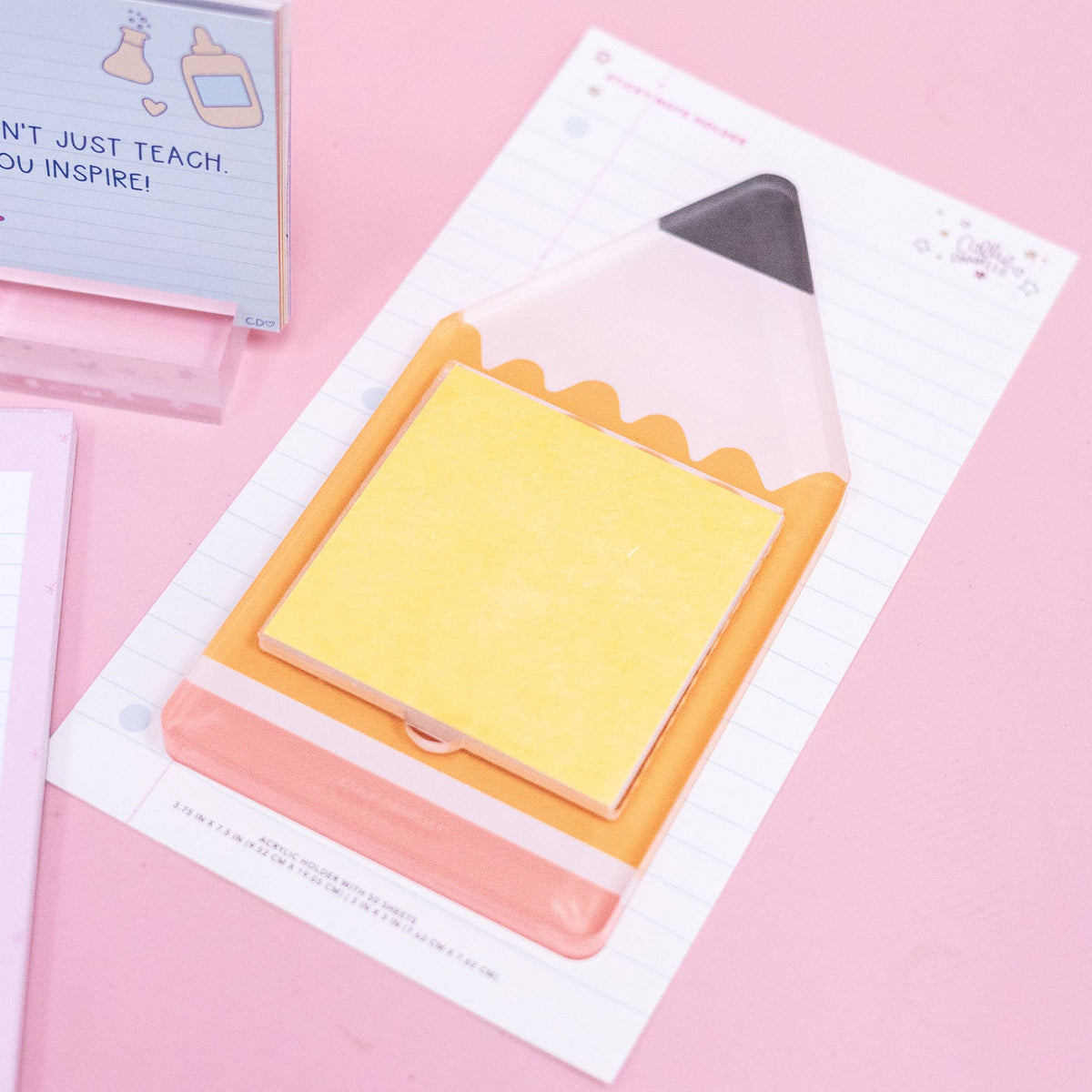 A pencil-shaped acrylic note holder by Callie Danielle, featuring yellow sticky notes on top and displayed on lined paper with a pink backdrop. Surrounded by other stationery essentials, the Callie Danielle - Pencil Sticky Note Holder makes an ideal back-to-school gift.