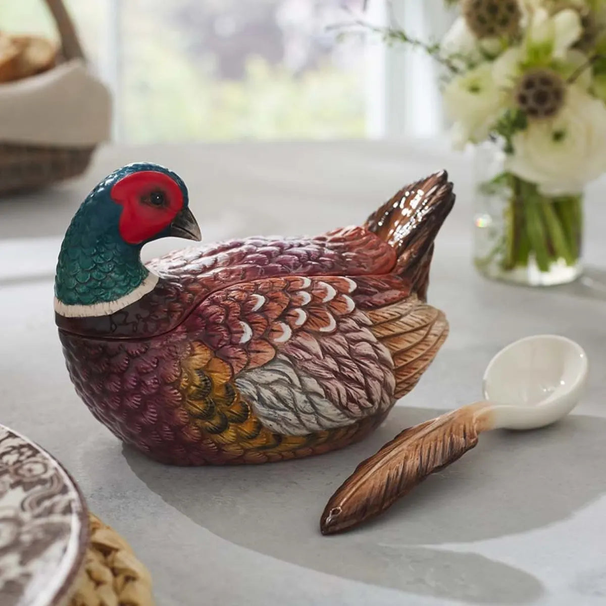 A Spode Woodland Pheasant Gravy Boat and Ladle from Spode, shaped like a colorful pheasant, sits on a table next to a bouquet of white flowers.
