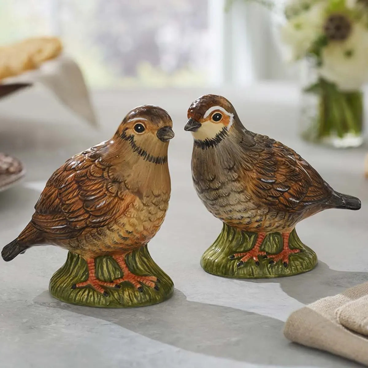 Two ceramic bird figurines modeled as quails, in brown and gray hues, rest on a table with a softly blurred background featuring flowers and bread. The Spode Woodland Quail Salt & Pepper set by Spode brings refined charm and elegance to any dining setting, making it an ideal piece of decorative tableware.