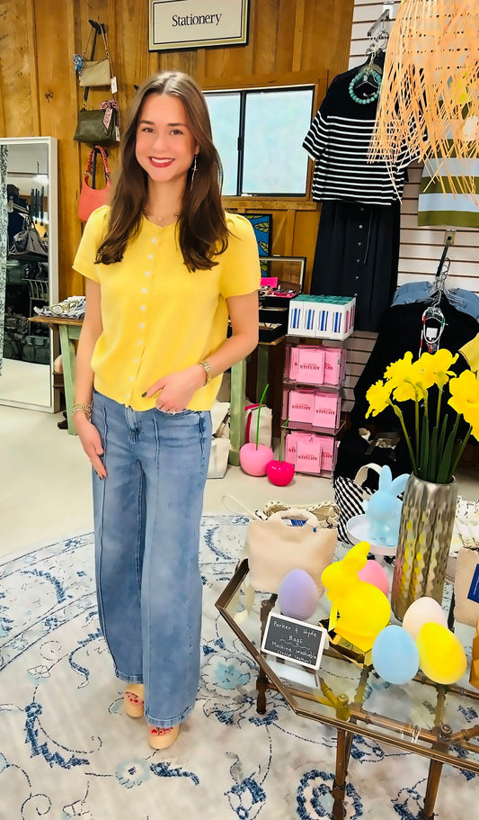 Wearing the &MERCI Short Sleeve Sweater Cardigan, a woman stands smiling in a boutique. Bright decor, daffodils, and colorful items are displayed on a glass table beside her, while shelves and racks with bags and clothes fill the background.