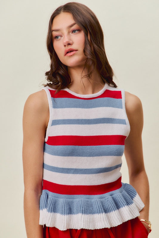 A woman with wavy brown hair wears the So Me 4th Of July Stripe Ruffle Bottom Sweater Knit Top by So Me. The sleeveless knit top features red, white, and blue horizontal stripes and a ruffled hem, creating a festive patriotic look perfect for a 4th of July party. She stands against a plain light background, gazing slightly to the side.