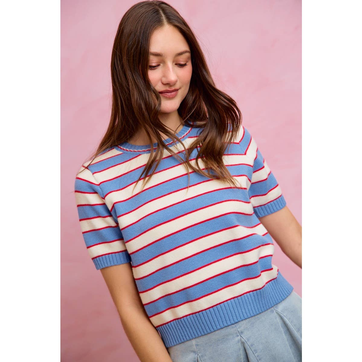 A woman with long brown hair wears the So Me STRIPE KNIT CREW NECK SHORT SLEEVES SWEATER TOP by So Me, featuring bold blue, white, and red stripes. She stands against a light pink background, looking down with a slight smile.