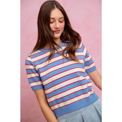A woman with long brown hair wears the So Me STRIPE KNIT CREW NECK SHORT SLEEVES SWEATER TOP by So Me, featuring bold blue, white, and red stripes. She stands against a light pink background, looking down with a slight smile.
