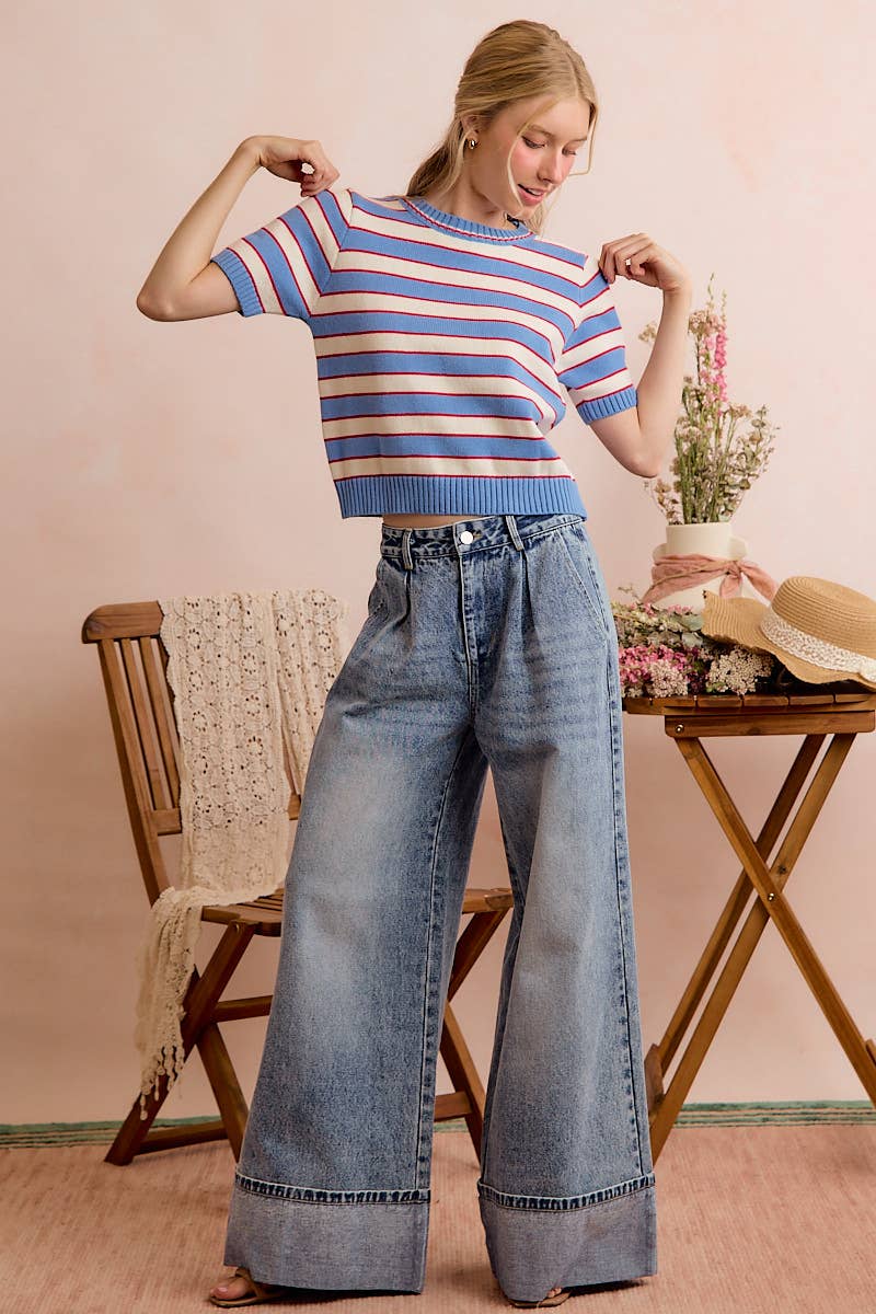 A woman poses indoors near a wooden chair draped with a lace shawl and a table decorated with flowers and straw hats, wearing the So Me STRIPE KNIT CREW NECK SHORT SLEEVES SWEATER TOP paired with wide-leg jeans. The background is light pink.