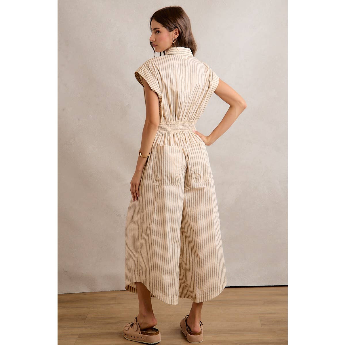 A woman stands on a wooden floor, facing away, wearing the So Me SLEEVELESS WIDE LEG STRIPE JUMPSUIT in beige and white stripes, paired with beige slip-on sandals. The scene is set against a plain light-colored wall.
